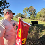 Man holding a red feed bag in a grassy field with a cow in the background